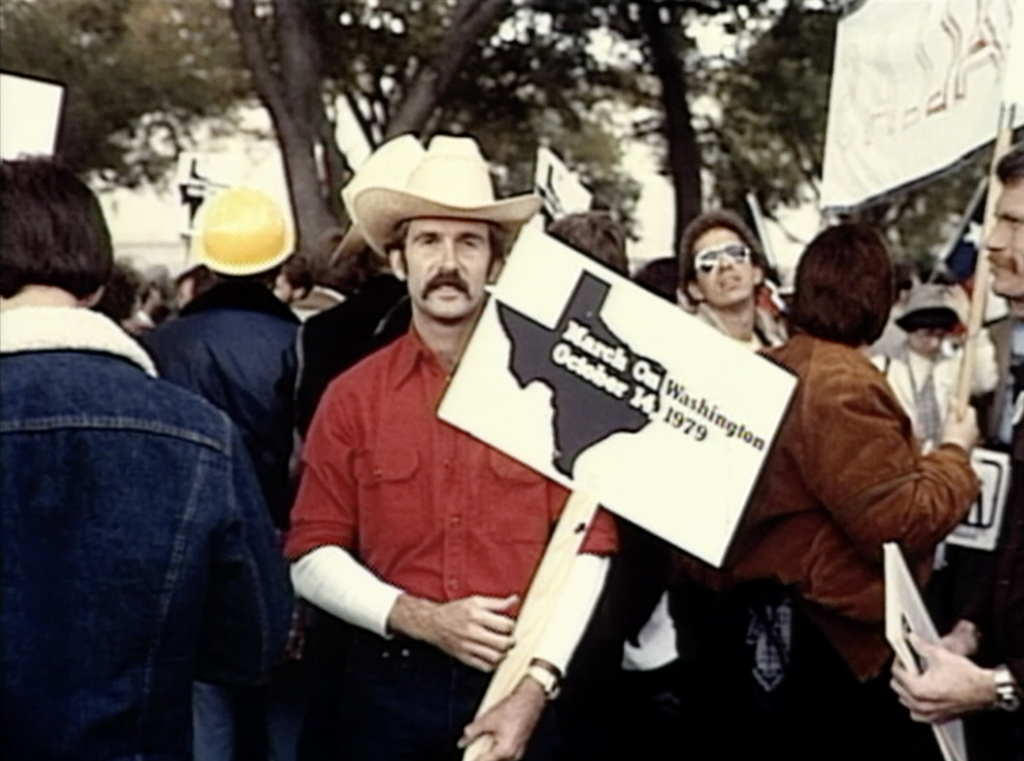 A man with a mustache, red shirt, and white cowboy hat smiles and holds a sign at a demonstration in D.C. in 1979.
